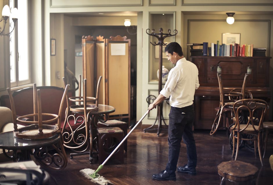 A professional cleaner wearing a face mask, gloves, and a white protective suit is performing deep cleaning or sanitisation on a white upholstered cushion using a handheld vacuum or cleaning device. T