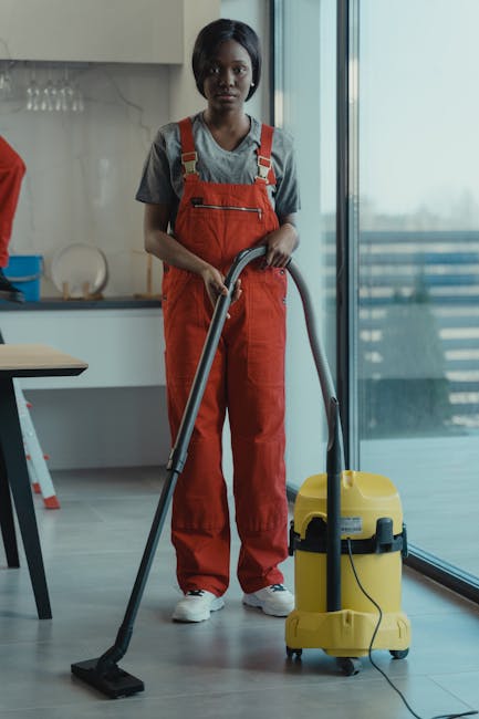 A female professional cleaner with dark hair, wearing a grey t-shirt and bright orange overalls, is operating a yellow commercial vacuum cleaner in a modern, well-lit room with large glass sliding doo